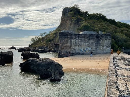 Plage rocheuse avec un ancien mur et une colline verdoyante en arrière-plan.
